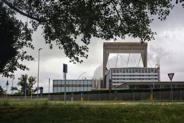 Ağaçlarla çerçevelenmiş çalışan bacaları ve yağmurlu bir günde yol olan bir fabrika.