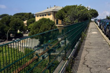 Güneşli bir günde, bir İtalyan şehrinde, otoyolun kenarında park halindeki arabalarla dolu bir ev.