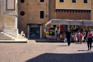 Güneşli bir günde bir İtalyan kasabasında bir tezgahın yanındaki meydanda oturan adam.