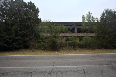 İtalya kırsalındaki terk edilmiş fabrika ağaçlarla çevrili ve yağmurdan önce bulutlu bir günde yolun karşı tarafından görülüyor.