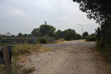 Bir köprünün yanındaki köy yolu ile bir ağacın çerçevelediği bulutlu bir günde toprak yol arasındaki kavşak.