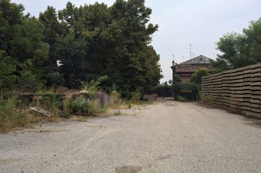 Bulutlu bir günde ağaçlarla çevrili bir grup ev ve kapıya açılan endüstriyel bir tesisin açıklıktaki çakıl yolu.