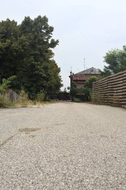 Bulutlu bir günde ağaçlarla çevrili bir grup ev ve kapıya açılan endüstriyel bir tesisin açıklıktaki çakıl yolu.