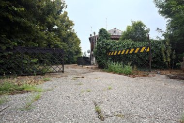 Bulutlu bir günde ağaçlarla çevrili bir grup ev ve kapıya açılan endüstriyel bir tesisin açıklıktaki çakıl yolu.