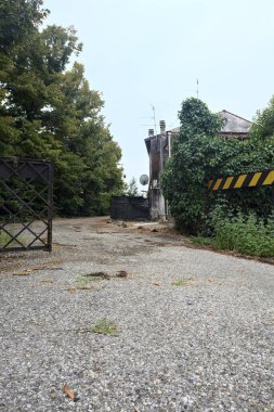 Bulutlu bir günde ağaçlarla çevrili bir grup ev ve kapıya açılan endüstriyel bir tesisin açıklıktaki çakıl yolu.