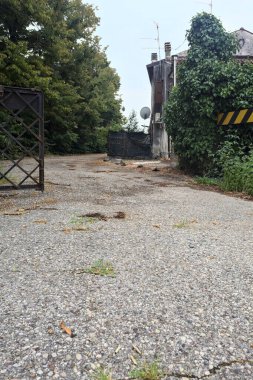 Bulutlu bir günde ağaçlarla çevrili bir grup ev ve kapıya açılan endüstriyel bir tesisin açıklıktaki çakıl yolu.