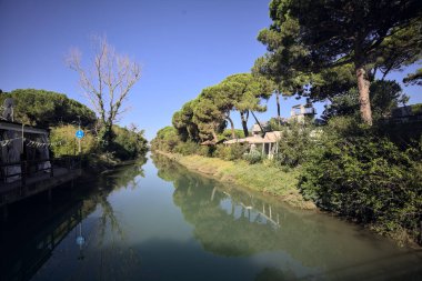Deniz çamları ve binalarla çevrili su akıntısı güneşli bir günde İtalyan kasabasında suya yansıyan yansımalarıyla.