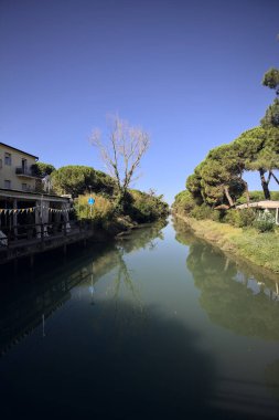 Deniz çamları ve binalarla çevrili su akıntısı güneşli bir günde İtalyan kasabasında suya yansıyan yansımalarıyla.