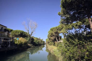 Deniz çamları ve binalarla çevrili su akıntısı güneşli bir günde İtalyan kasabasında suya yansıyan yansımalarıyla.
