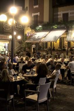Meydanda bir restoran dolusu insan ve geceleri deniz kıyısındaki bir İtalyan kasabasına girişi var.
