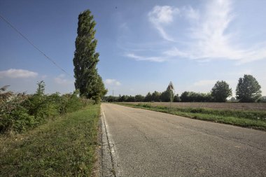 İtalya kırsalında güneşli bir günde tarlaların yanında seyrek ağaçlarla çevrili bir köy yolu.
