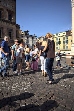 Bir İtalyan kasabasının meydanı. Yakınlardaki bir caddeden insanların geçtiği görülüyor.