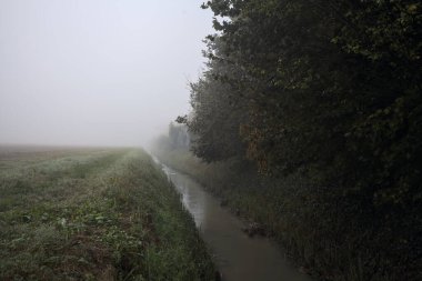 İtalya kırsalında sisli bir günde, bir blok ev ve bir tarla ile çevrili bir hendek.