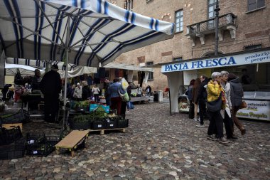 Bir italyan kasabasında kaldırımlı bir meydanda tezgahlar ve insanlar onları tarıyor.