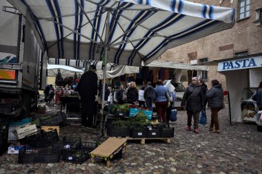 Bir italyan kasabasında kaldırımlı bir meydanda tezgahlar ve insanlar onları tarıyor.