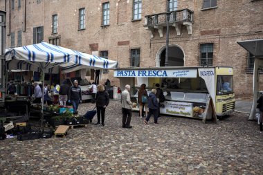 Bir italyan kasabasında kaldırımlı bir meydanda tezgahlar ve insanlar onları tarıyor.