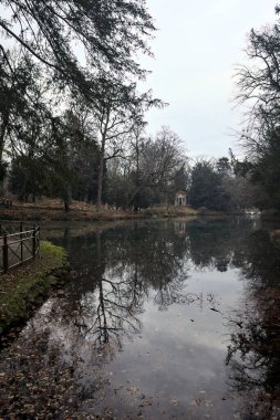 Kıyısında neoklasik bir tapınağı olan bir parkta gölet ve sonbaharda bulutlu bir günde ağaçların yansıması.