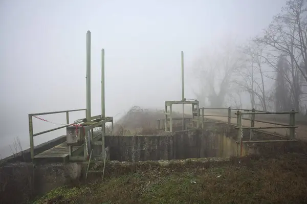 Weir by the edge of a dirt path on a foggy day