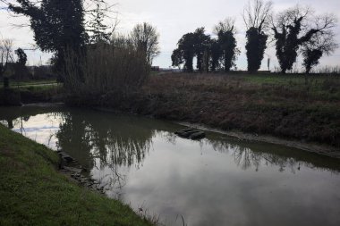 İtalya 'nın kırsal kesimlerinde bulutlu bir günde sudaki ağaçlarla dolu küçük bir köprünün altından geçen dere.