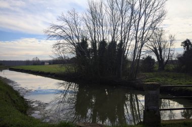 İtalya 'nın kırsal kesimlerinde bulutlu bir günde sudaki ağaçlarla dolu küçük bir köprünün altından geçen dere.
