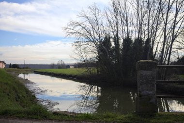 İtalya 'nın kırsal kesimlerinde bulutlu bir günde sudaki ağaçlarla dolu küçük bir köprünün altından geçen dere.