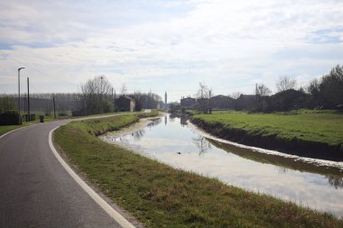 Bulutlu bir gökyüzü ve uzak bir çan kulesi olan bir köyün yanındaki su deresi İtalya kırsalındaki suya yansıyor.