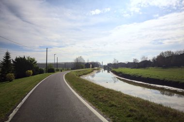Bulutlu bir gökyüzü ve uzak bir çan kulesi olan bir köyün yanındaki su deresi İtalya kırsalındaki suya yansıyor.