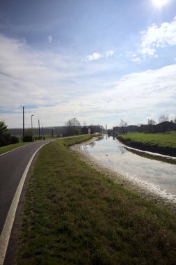 Bulutlu bir gökyüzü ve uzak bir çan kulesi olan bir köyün yanındaki su deresi İtalya kırsalındaki suya yansıyor.