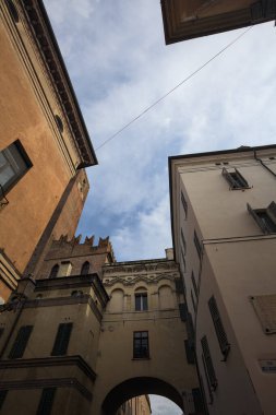 Tarihi binaların cepheleri ve arka plan olarak kısmen bulutlu gökyüzü ile ayakta duran tuğla bir kule.