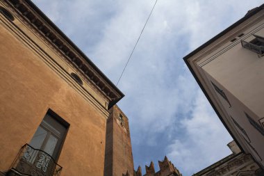 Tarihi binaların cepheleri ve arka plan olarak kısmen bulutlu gökyüzü ile ayakta duran tuğla bir kule.