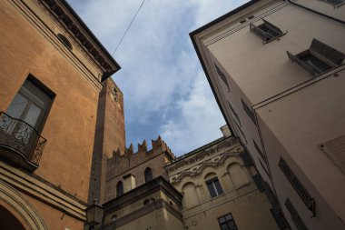 Tarihi binaların cepheleri ve arka plan olarak kısmen bulutlu gökyüzü ile ayakta duran tuğla bir kule.