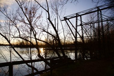 Gün batımında bir parkta nehrin kenarında metalik bir yapının altından geçen patika.