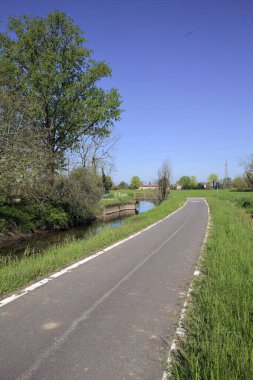 İtalya kırsalında güneşli bir günde üzerinden küçük beton bir köprü geçen bir su deresinin yanındaki patikada.