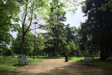 Güneşli bir günde, bir İtalyan kasabasının parkındaki gölgede kısmen ahşap bankların olduğu patika.