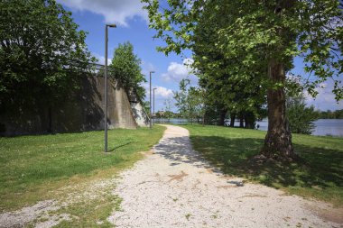 Güneşli bir günde, gölün kıyısındaki beton set ve ağaçlar arasındaki toprak izi.