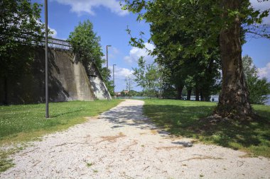 Güneşli bir günde, gölün kıyısındaki beton set ve ağaçlar arasındaki toprak izi.