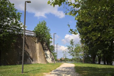Güneşli bir günde, gölün kıyısındaki beton set ve ağaçlar arasındaki toprak izi.