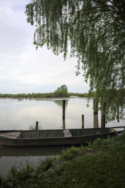 Nehir kıyısında bir söğüt ağacının yanında demirli bir tekne ve suya düşen bulutlu bir gökyüzü.