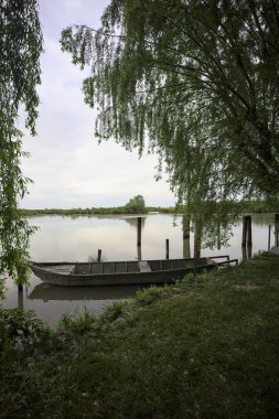Nehir kıyısında bir söğüt ağacının yanında demirli bir tekne ve suya düşen bulutlu bir gökyüzü.
