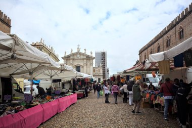 Mantova, İtalya - 8. Mart 2025 - Arka planda bir kilise olan bir sarayın ve insanların market tezgahlarına göz attığı bir meydanın yanındaki kaldırımlı meydan