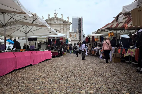 Mantova, İtalya - 8. Mart 2025 - Arka planda bir kilise olan bir sarayın ve insanların market tezgahlarına göz attığı bir meydanın yanındaki kaldırımlı meydan
