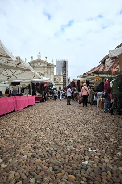Mantova, İtalya - 8. Mart 2025 - Arka planda bir kilise olan bir sarayın ve insanların market tezgahlarına göz attığı bir meydanın yanındaki kaldırımlı meydan