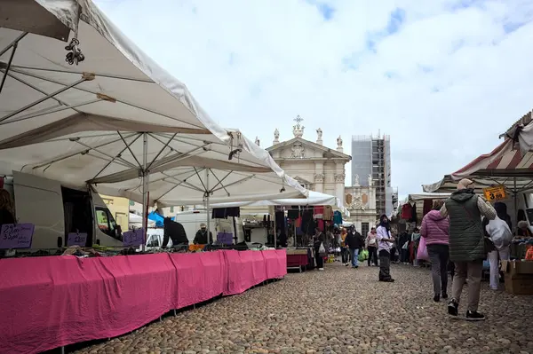 Mantova, İtalya - 8. Mart 2025 - Arka planda bir kilise olan bir sarayın ve insanların market tezgahlarına göz attığı bir meydanın yanındaki kaldırımlı meydan