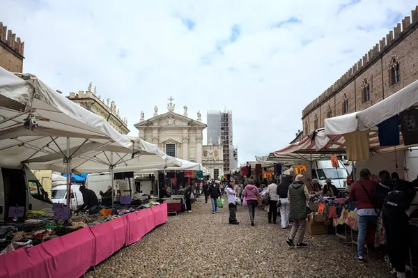 Mantova, İtalya - 8. Mart 2025 - Arka planda bir kilise olan bir sarayın ve insanların market tezgahlarına göz attığı bir meydanın yanındaki kaldırımlı meydan