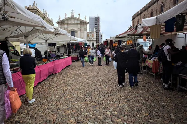 Mantova, İtalya - 8. Mart 2025 - Arka planda bir kilise olan bir sarayın ve insanların market tezgahlarına göz attığı bir meydanın yanındaki kaldırımlı meydan