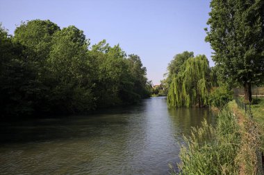 İtalya kırsalında güneşli bir günde, kıyılarında salkım söğüdü olan bir parkın içinden geçen nehir.