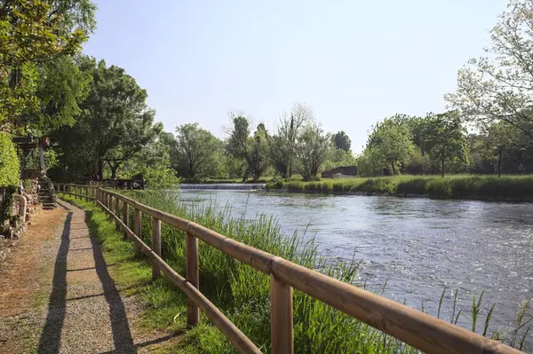 İtalya kırsalında güneşli bir günde nehir kıyısında ahşap bir demiryolu ile çevrili toprak ve dar bir patika.