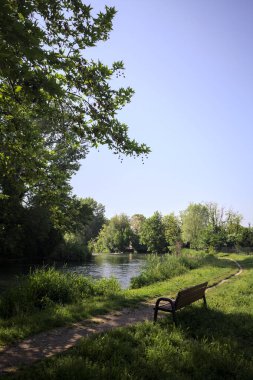 Tahta bank, kısmen gölgede, nehir kıyısında, İtalya kırsalındaki bir parkın yanında.