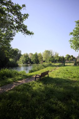 Tahta bank, kısmen gölgede, nehir kıyısında, İtalya kırsalındaki bir parkın yanında.
