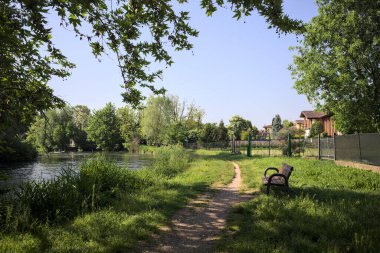 Tahta bank, kısmen gölgede, nehir kıyısında, İtalya kırsalındaki bir parkın yanında.